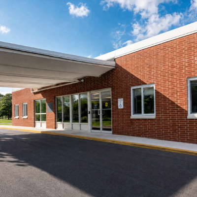 A brick rehabilitation center building with large glass doors and windows, a white overhang, and a parking lot in front under a partly cloudy blue sky.