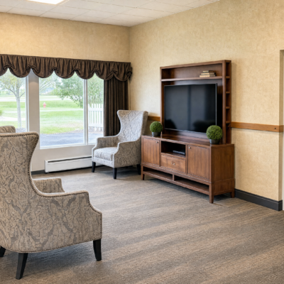 A cozy living room with two armchairs, a large window with dark curtains, and a wooden entertainment center with a flat-screen TV. The room has beige walls and carpeted floor.