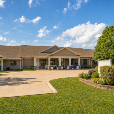 A single-story rehabilitation center building with a gable roof, beige exterior walls, and a covered entrance supported by white columns. The building is surrounded by a well-maintained lawn, colorful flower beds, and a large paved driveway under a blue sky with scattered clouds.
