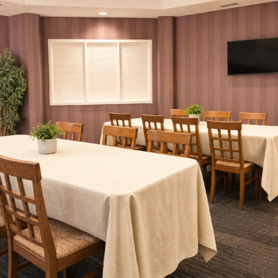 A conference room with two long tables covered in white tablecloths, surrounded by wooden chairs, a potted plant on each table, a window with closed blinds, and a wall-mounted TV on a striped purple wall.