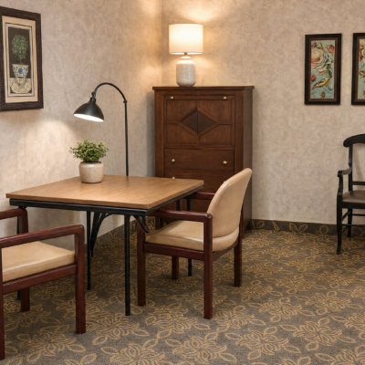 A cozy room with a wooden table and two cushioned chairs. On the table, there is a small potted plant and a black adjustable desk lamp. Against the wall, there is a wooden cabinet with a white table lamp on top. The walls are decorated with framed artwork, and the floor has a patterned carpet.